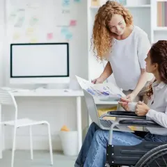 Kobieta na wózku inwalidzkim omawia wykresy z koleżanką. W miejscu pracy, gdzie stopnie niepełnosprawności nie ograniczają, a wręcz dają pewne przywileje, obie panie współpracują.