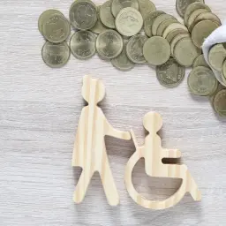 Drewniane figurki osoby pomagającej osobie na wózku inwalidzkim i monety. Czy zasiłek pielęgnacyjny wlicza się do dochodu?