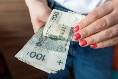 Dłonie trzymające stos banknotów 100 zł. Porównanie zasiłku stałego a renty socjalnej.