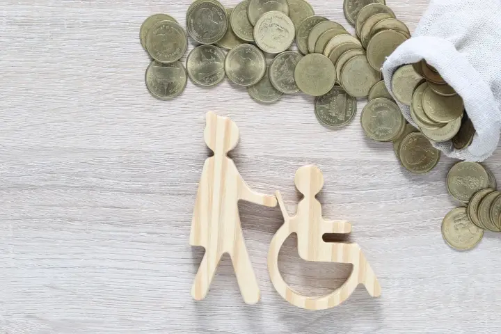 Drewniane figurki osoby pomagającej osobie na wózku inwalidzkim i monety. Czy zasiłek pielęgnacyjny wlicza się do dochodu?