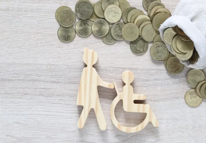 Drewniane figurki osoby pomagającej osobie na wózku inwalidzkim i monety. Czy zasiłek pielęgnacyjny wlicza się do dochodu?