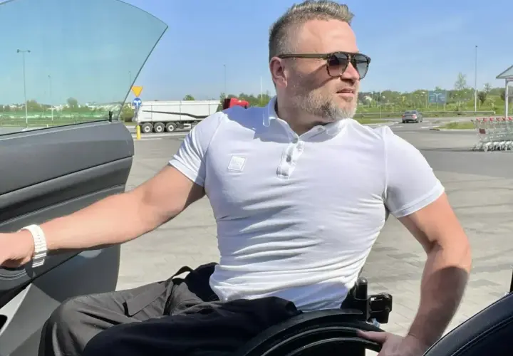 Mężczyzna na wózku inwalidzkim przy otwartych drzwiach samochodu na parkingu.