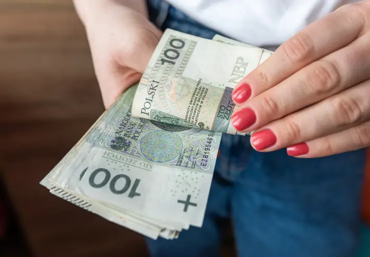 Dłonie trzymające stos banknotów 100 zł. Porównanie zasiłku stałego a renty socjalnej.